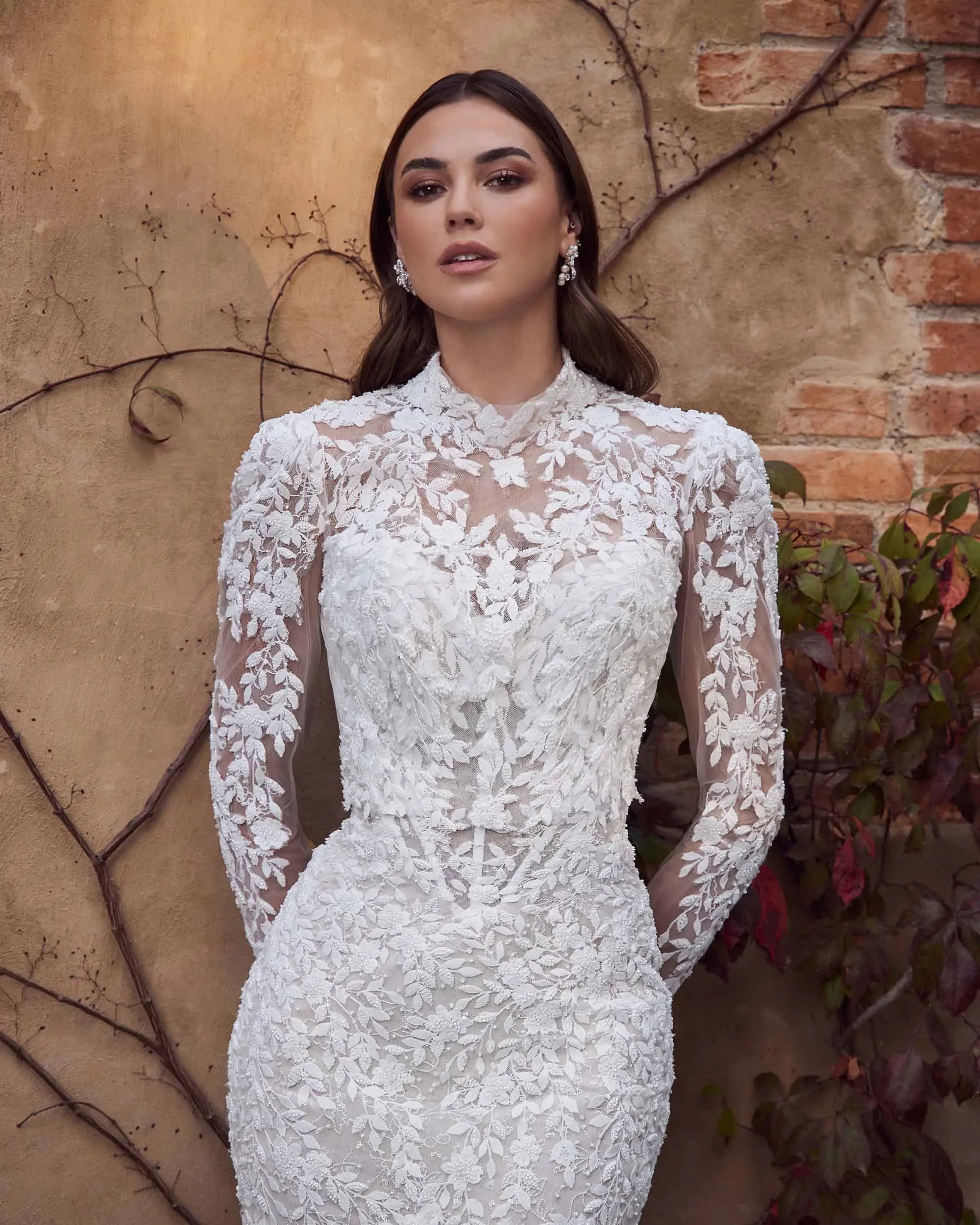 A woman in an elegant, lace-patterned white wedding gown stands against a rustic wall with vine details, exuding sophistication and grace.