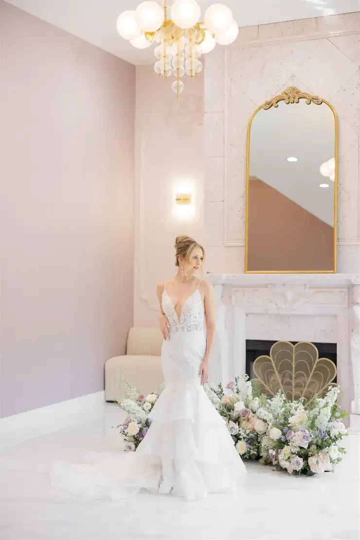A bride in a white wedding gown stands elegantly in a beautifully decorated room with a floral arrangement.