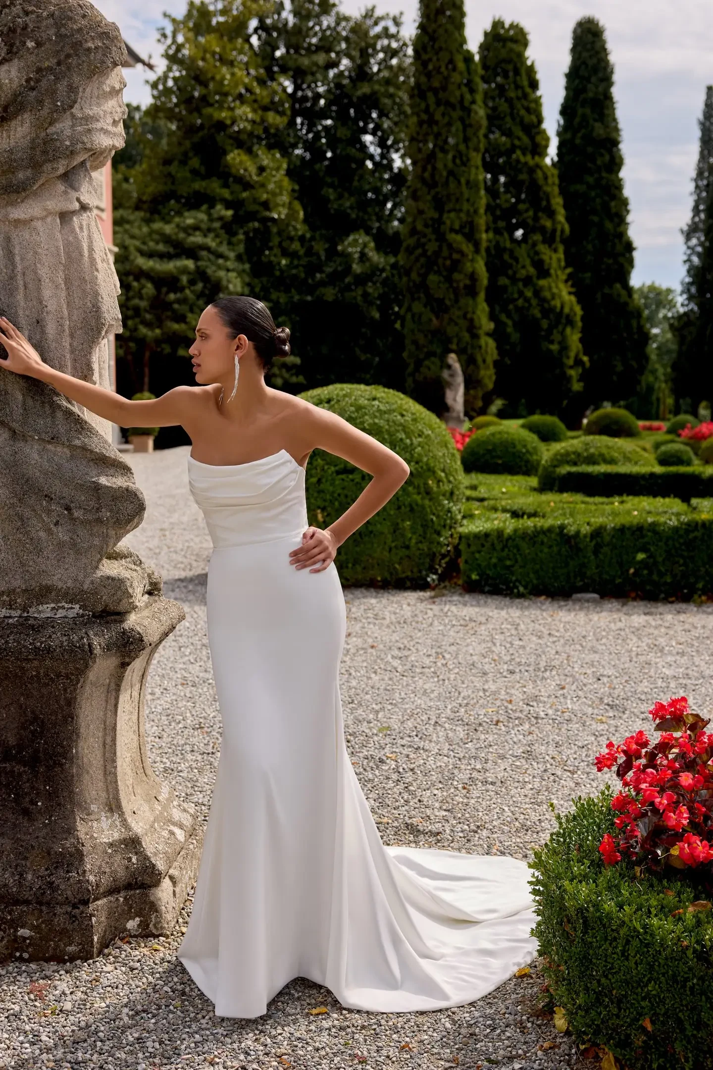 A woman in a strapless white gown standing near a stone structure in a garden with manicured hedges and flowers.