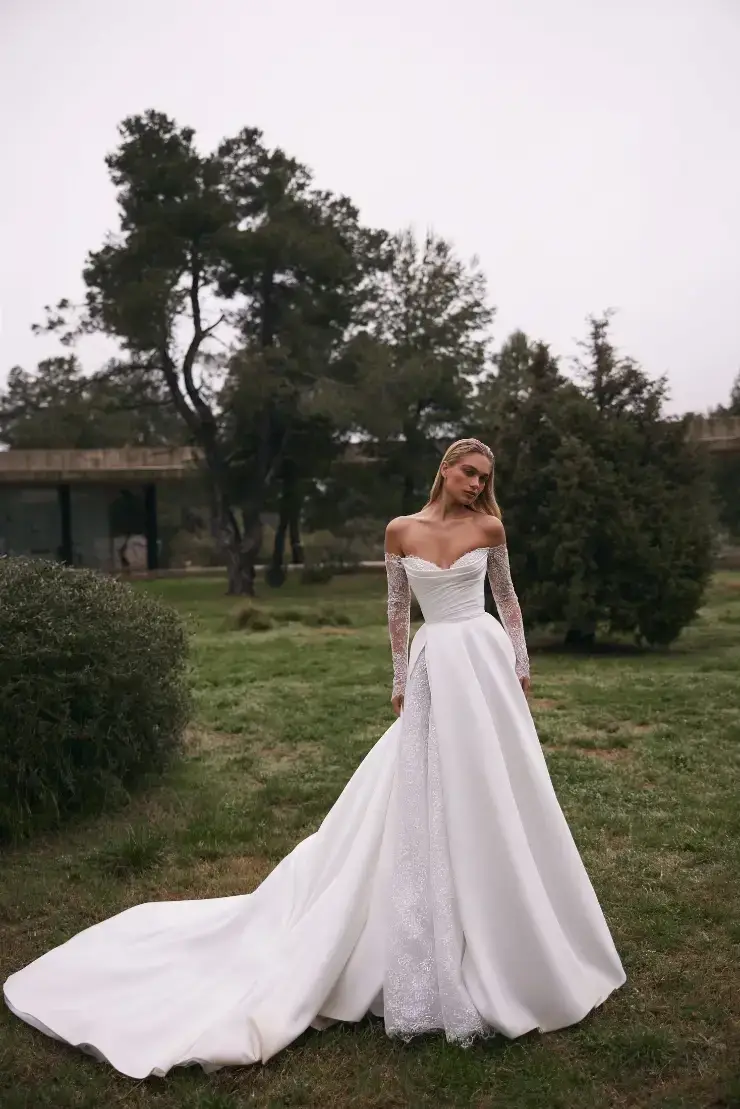 A woman in a white wedding dress with long sleeves, standing in a grassy area surrounded by trees.