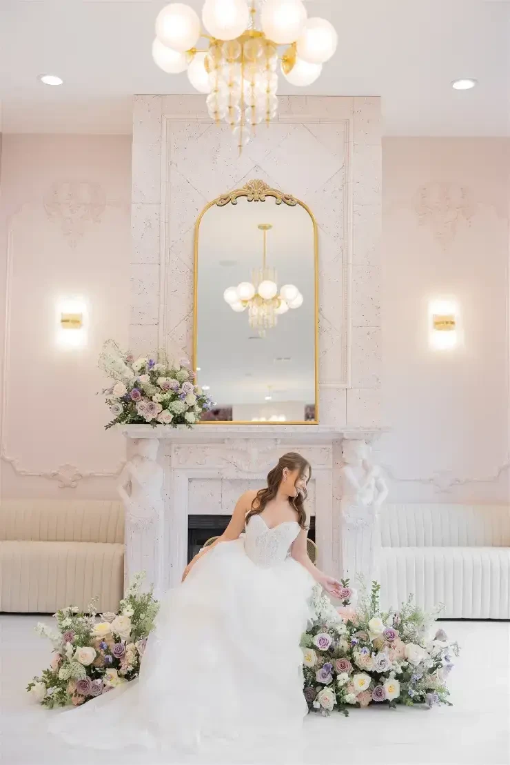 A bride in a white gown posing in front of a fireplace adorned with flowers.