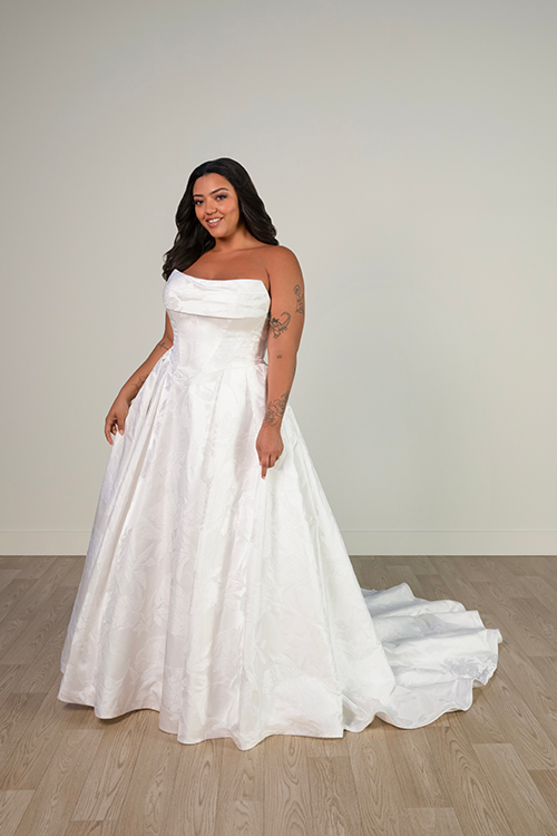 A woman wearing a white wedding dress with a fitted bodice and flowing skirt, standing in a bright, minimalistic room.
