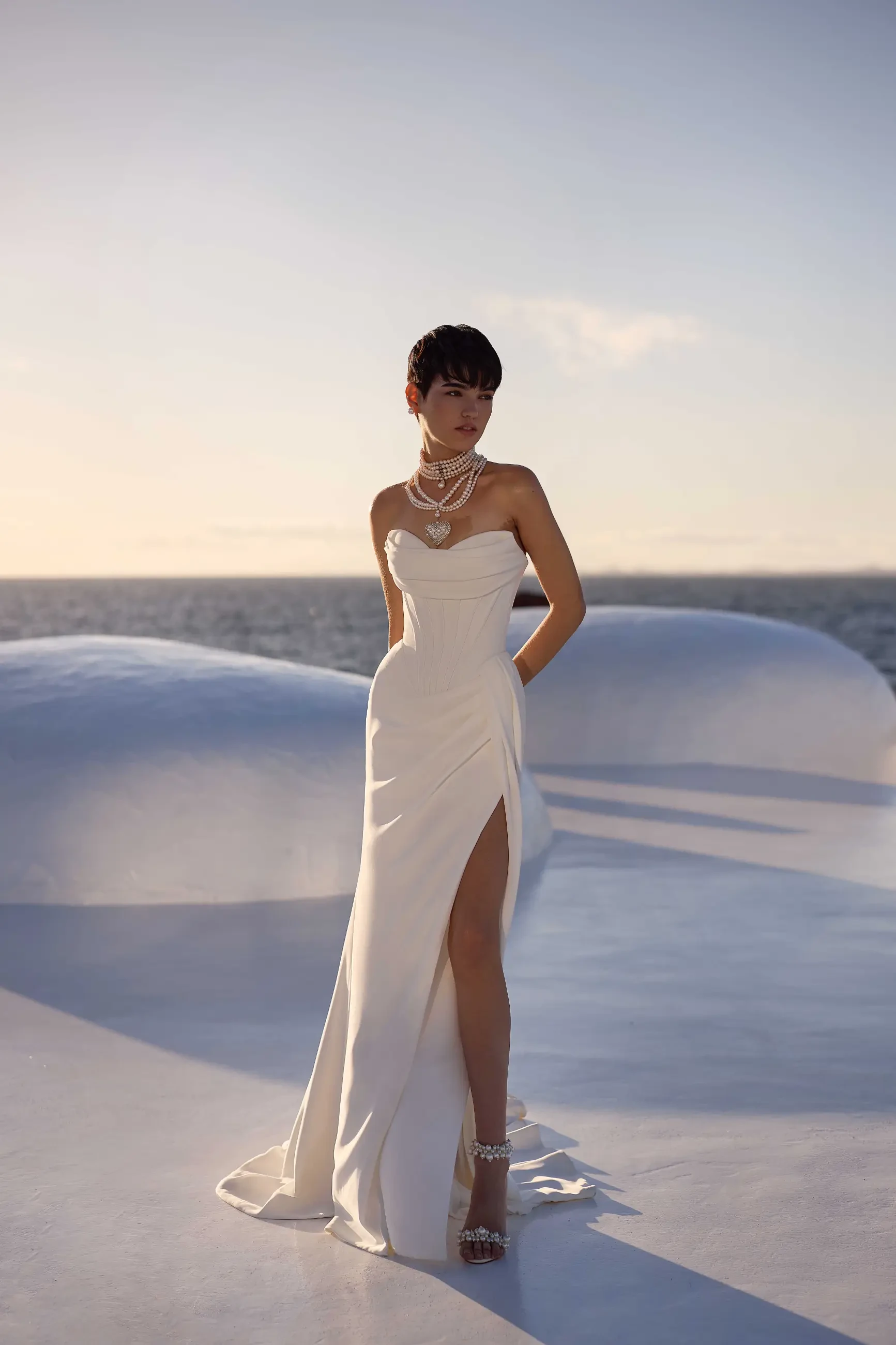 A woman in an elegant white strapless gown stands outdoors, with the ocean and a soft sunset in the background. She wears layered necklaces, evoking a serene and sophisticated mood.