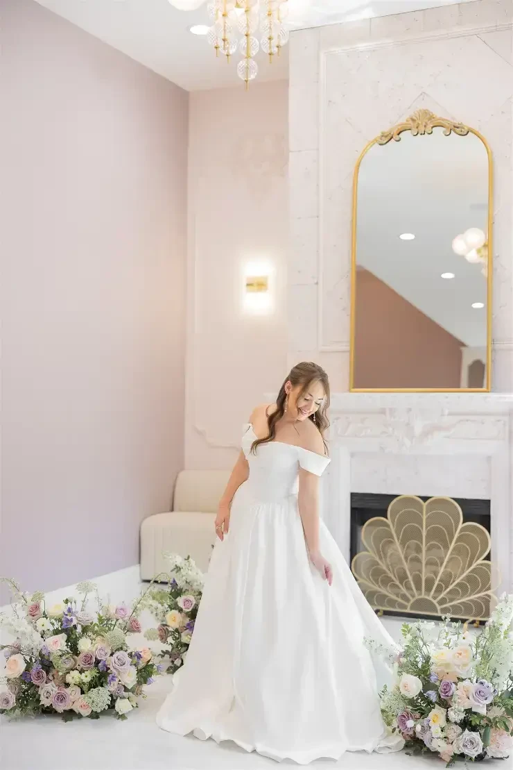 A bride in a white off-shoulder wedding dress stands gracefully in a beautifully decorated room with floral arrangements.