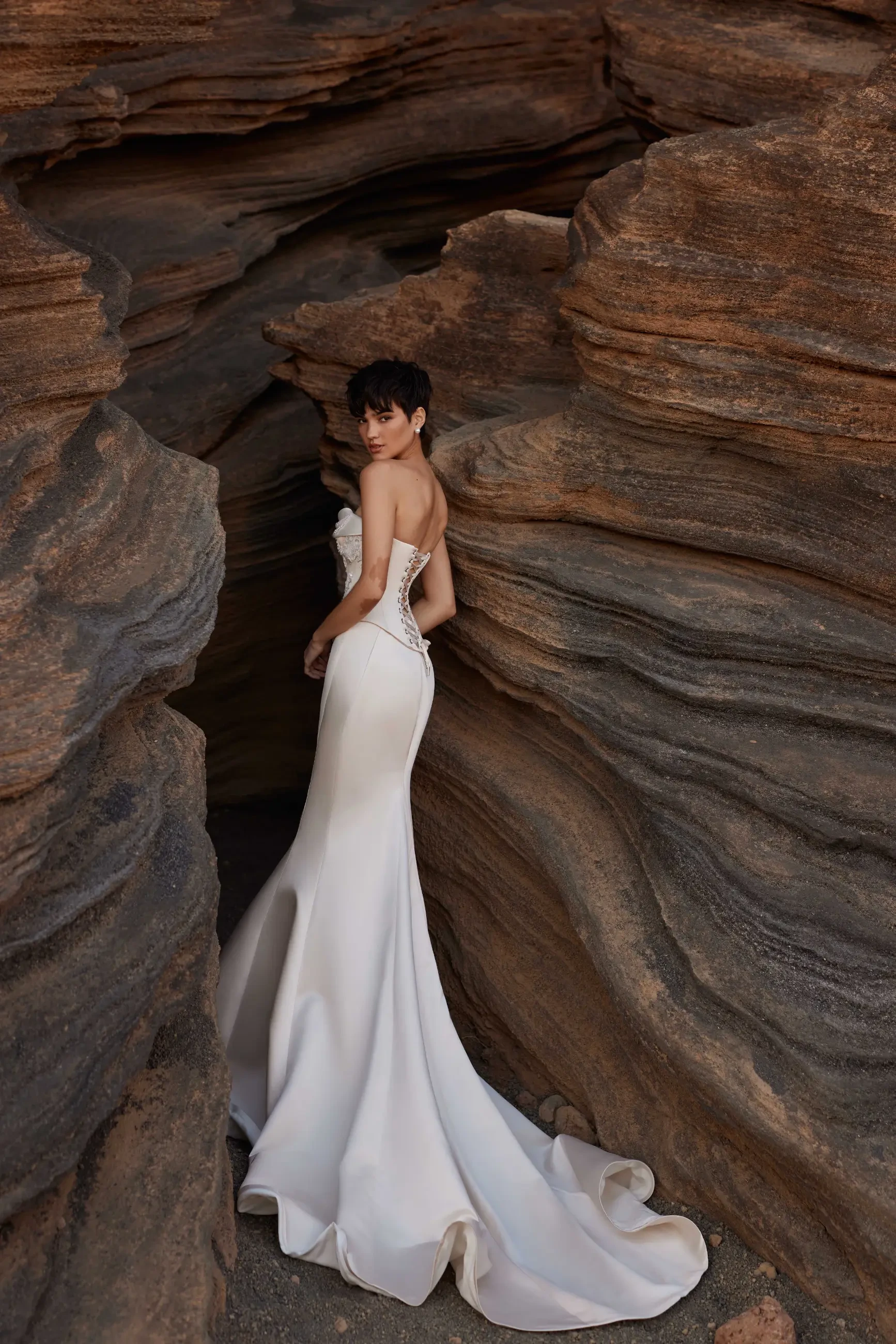 A woman in a strapless white gown stands gracefully between layered rock formations, gazing back. The scene conveys elegance and natural beauty.