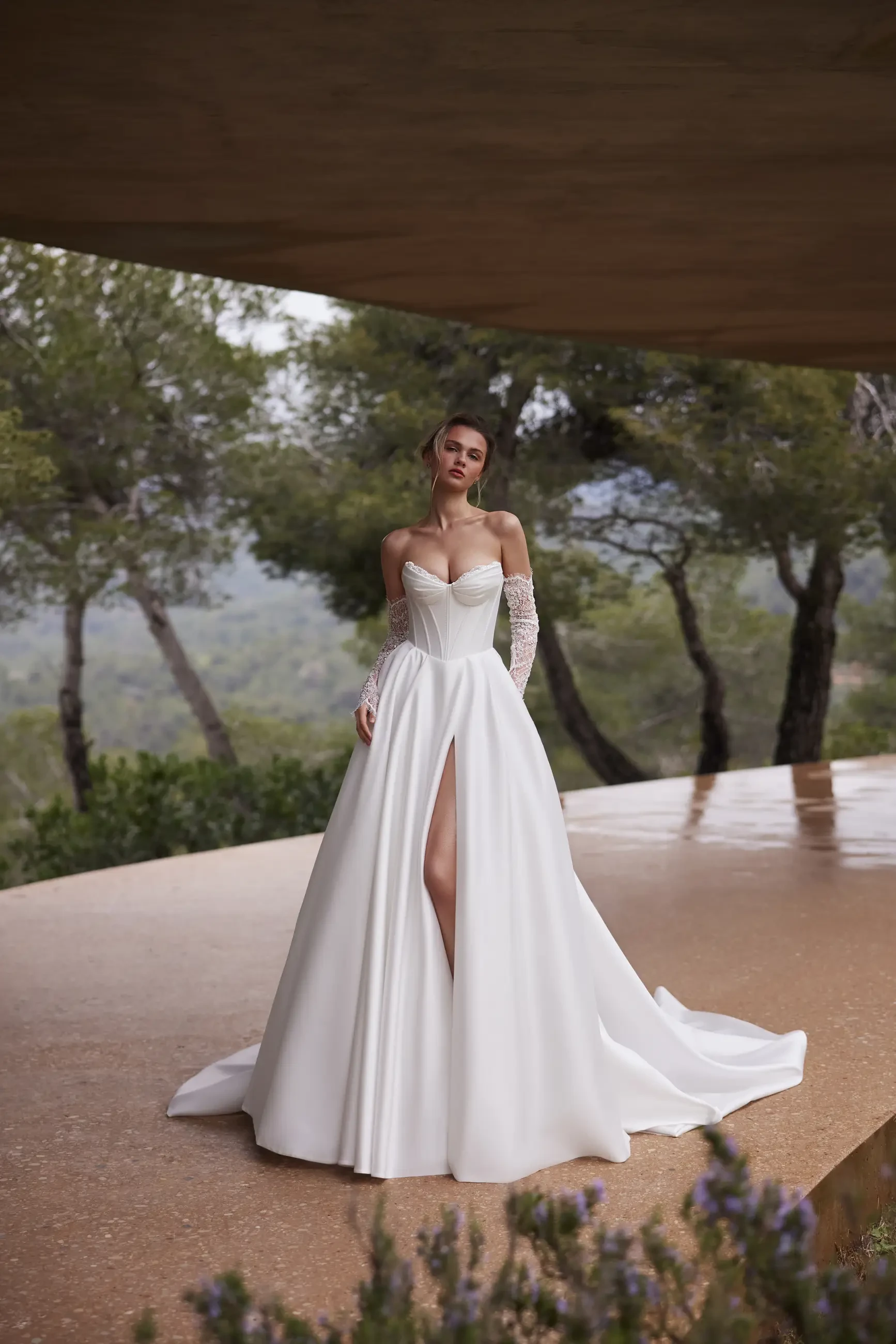 Bride in an elegant, strapless white gown with a high slit and lace gloves stands on a terrace. Lush trees in the background, exuding a serene, romantic feel.