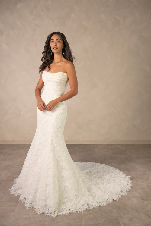 A woman wearing a strapless white wedding dress with a fitted bodice and a flared lace train, standing against a neutral background.