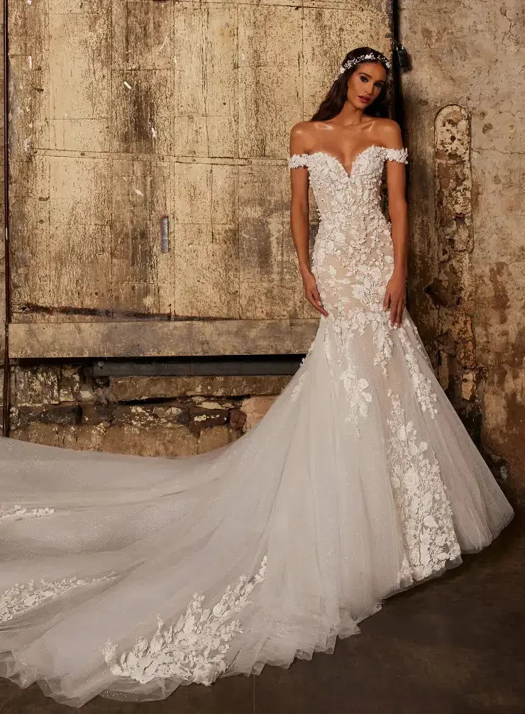 A model wearing a stunning off-the-shoulder wedding dress with intricate lace detailing and a long, flowing train, posing in an industrial setting.