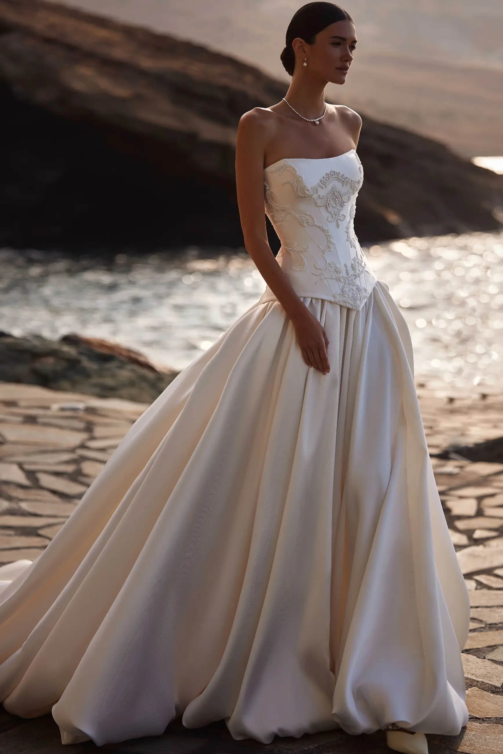 Bride in a flowing strapless gown stands on a rocky coastline. The setting sun casts a warm glow, highlighting the dress's intricate lace details. Romantic ambiance.