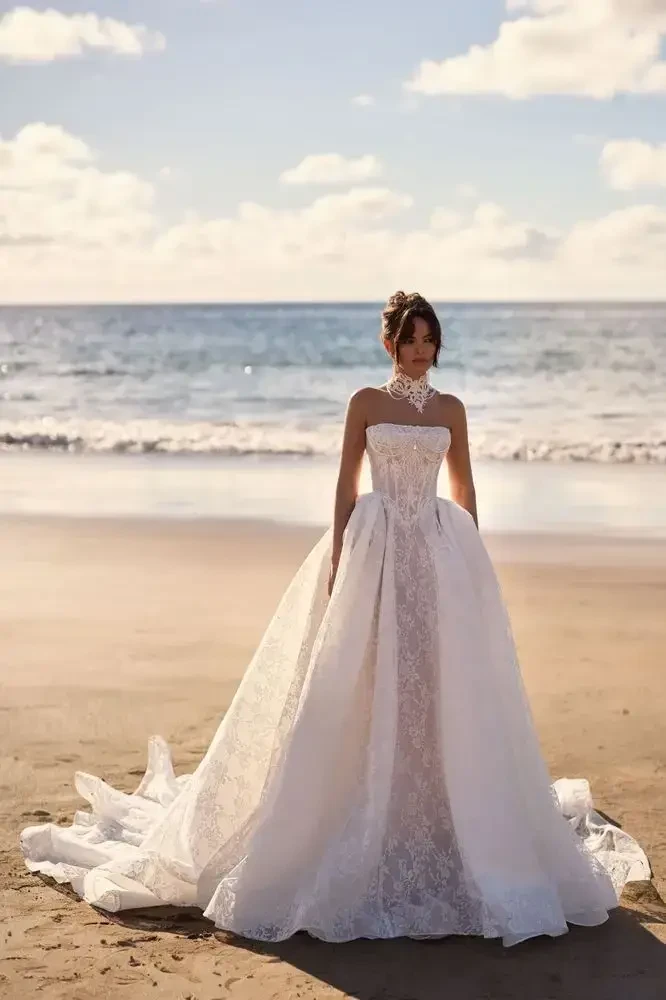 A woman in a wedding dress stands on a beach with the ocean in the background.