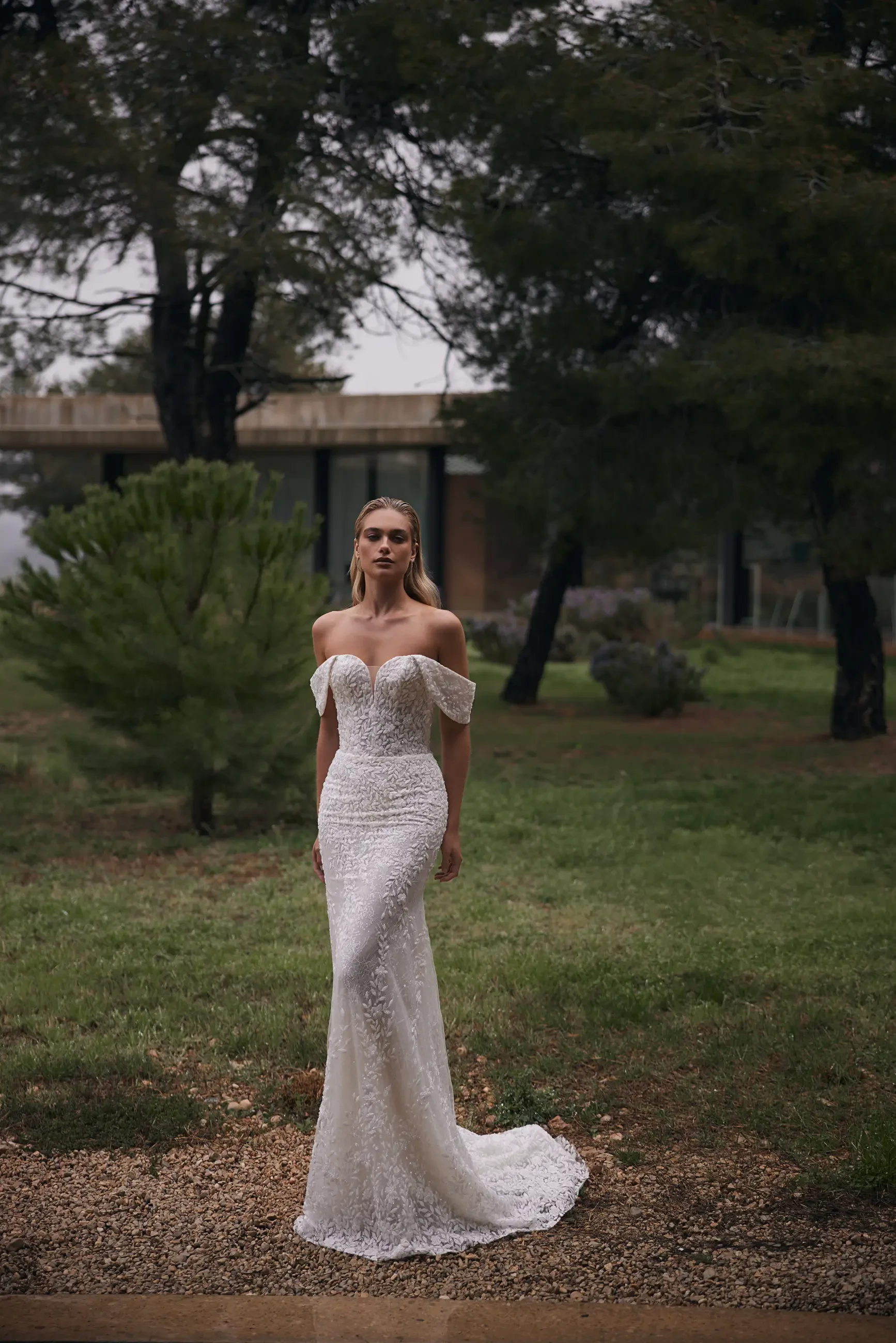A bride stands in a lush garden wearing an elegant off-shoulder lace gown. The overcast sky and trees create a serene, romantic atmosphere.