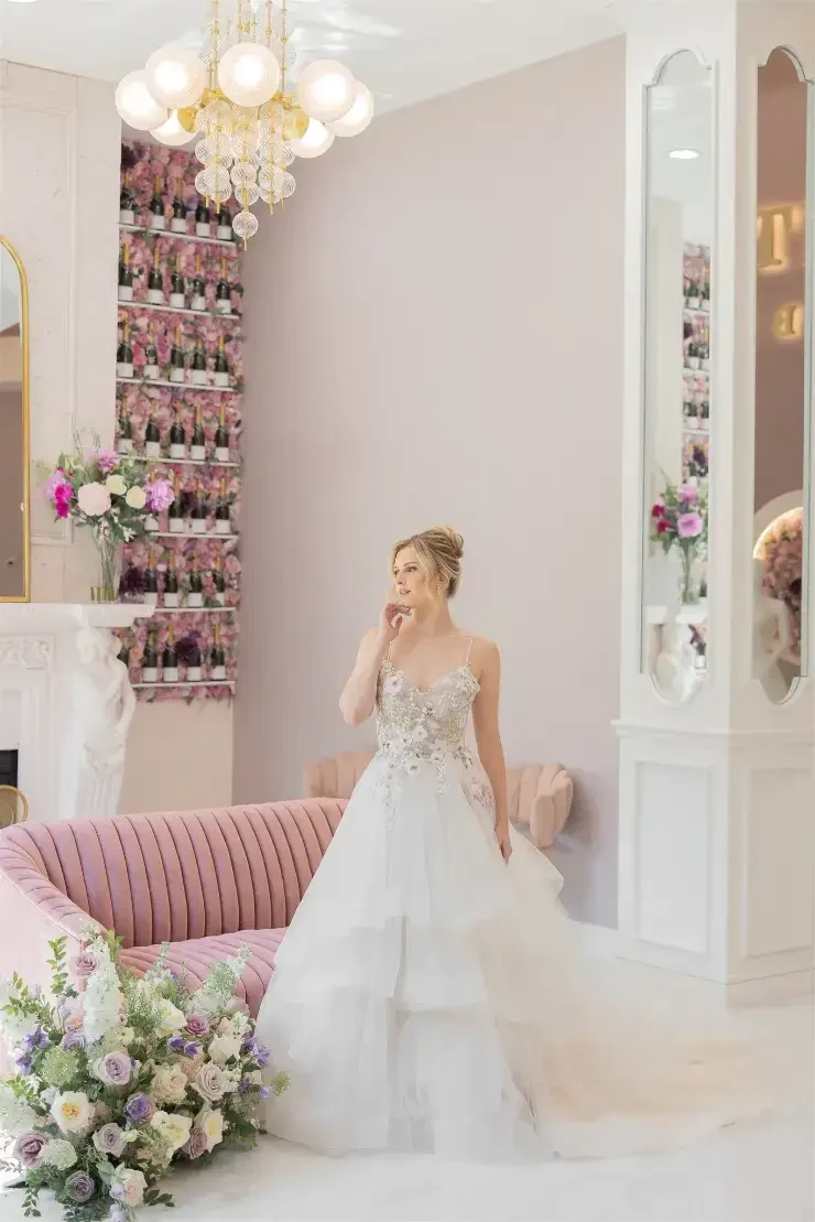 A bride in a decorative wedding dress stands in a stylish interior, next to a pink sofa and a bouquet of flowers.