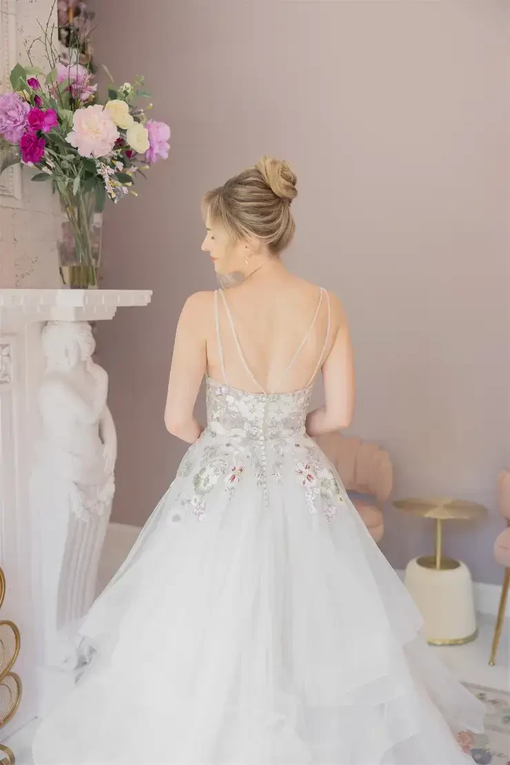 A bride in a decorative wedding dress stands in a stylish interior, next to a pink sofa and a bouquet of flowers.