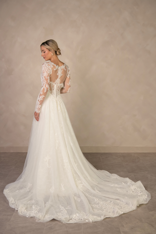 A woman poses elegantly in a long-sleeved wedding dress with lace details and a flowing train, standing against a neutral background.