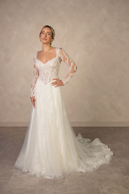 A woman poses elegantly in a long-sleeved wedding dress with lace details and a flowing train, standing against a neutral background.
