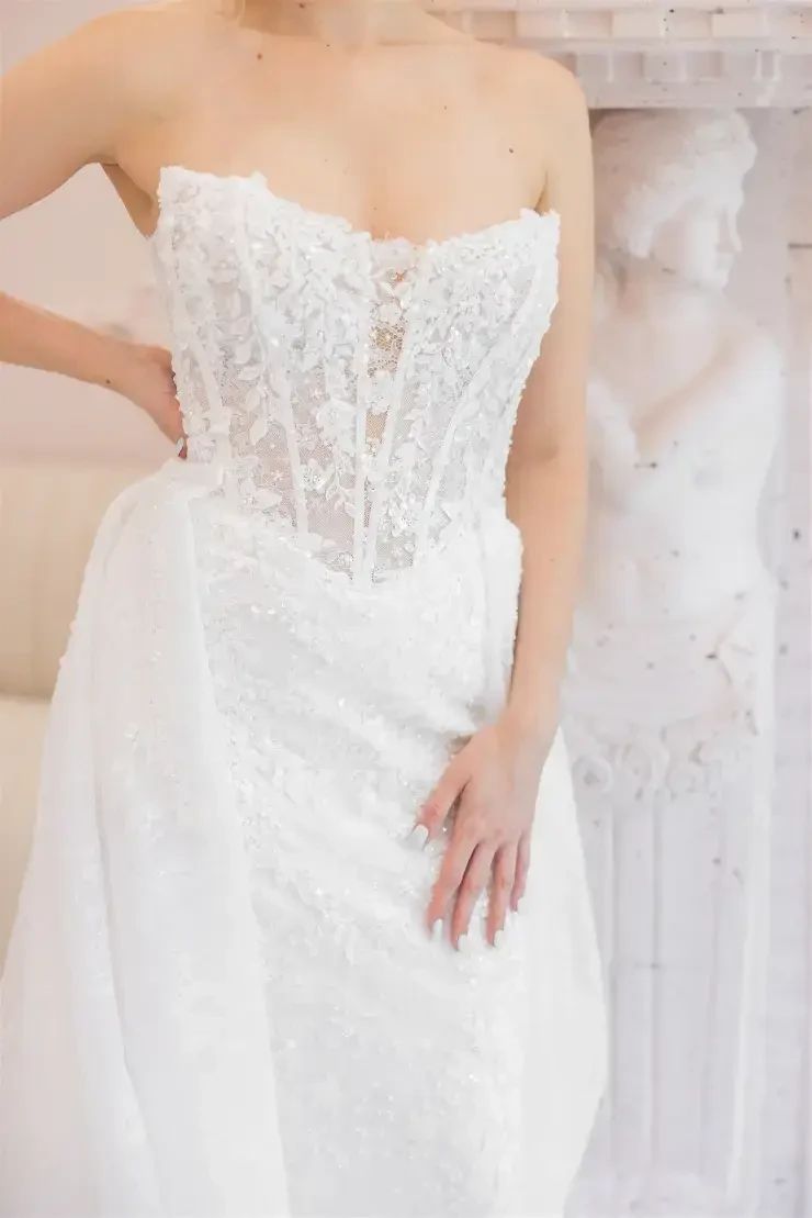 A close-up of a person wearing a beautiful white wedding dress with intricate lace details, standing in a bright, elegant setting.