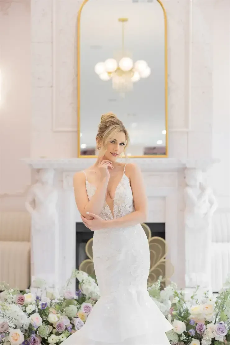 A bride in a fitted white wedding dress stands elegantly in a beautifully decorated room with a floral arrangement and a large mirror.